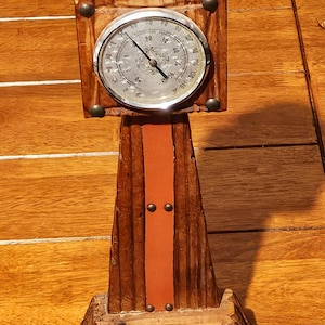 May include: A vintage wooden barometer with a circular gauge, featuring a silver face and black markings. The wooden frame is intricately carved and stained a warm brown, with a vertical orange leather strip. Decorative metal studs add detail.