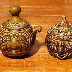 May include: Two olive-green glazed ceramic jars with lids. The left jar features a rounded lid with a knob, side handles, and the word Onions. The right jar has a textured lid with vegetable motifs and the word CHUTNEY. Both jars feature faces.