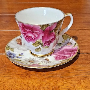 May include: A white porcelain teacup and matching saucer featuring a vibrant floral pattern of large pink roses and smaller white and blue daisies. Both the cup and saucer are finished with a delicate gold trim along the scalloped edges.