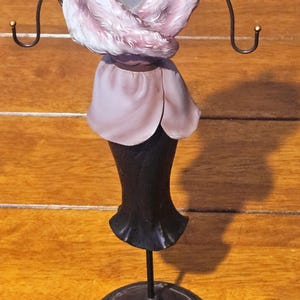 Könnte beinhalten: Dekorativer Schmuckständer in Form einer Schneiderpuppe. Der Ständer hat einen schwarzen Sockel und Stiel, einen mauvefarbenen Oberteil und einen Kunstfellschal. Der Ständer hat gebogene Arme zum Aufhängen von Schmuck.