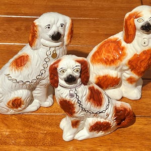 May include: Three ceramic dog figurines featuring white coats with orange-brown patches. Each dog has painted facial features, including dark noses and eyes. Two of the dogs wear painted collars with attached chains and small circular tags.