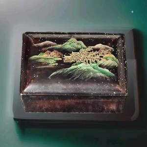 May include: A black lacquered box with a gold and green landscape scene painted on the lid. The scene features mountains, trees, and a small building. The box is decorated with gold glitter.