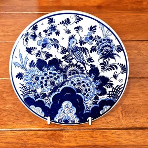 Op de afbeelding: Een ronde, witte keramische plaat met een blauw bloemen- en vogelontwerp. De plaat heeft een gedetailleerd patroon van bloemen, bladeren en een zittende vogel, allemaal in blauwtinten. De plaat wordt gepresenteerd op een houten oppervlak.
