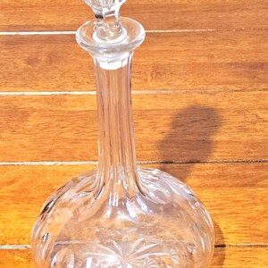 Puede incluir: Un decantador de vidrio transparente con un tapón esférico y un cuello largo y delgado. La base del decantador es redondeada y está decorada con una serie de cortes verticales y un patrón de estrella grabado en la parte inferior del vidrio.
