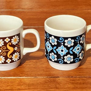 May include: Two white ceramic mugs with floral patterns. One has a brown and yellow daisy design, the other a blue and white floral pattern on black. Both mugs have white handles. Ideal for tea or coffee.