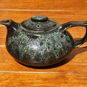 May include: A ceramic teapot featuring a dark green and black mottled glaze with a glossy finish. The teapot has a rounded body, a curved spout, a sturdy handle, and a matching lid with a small knob and a steam vent hole on the top surface.