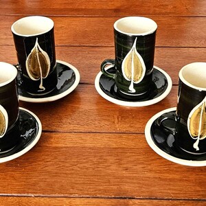 May include: A set of four tall ceramic cups and matching saucers featuring a dark green glaze with a prominent gold and white leaf motif on the front of each cup. The interior of each cup is a light cream colour, and the saucers have a cream rim.