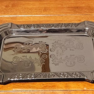 Puede incluir: Una bandeja rectangular plateada con asas ornamentadas y un diseño floral. La bandeja tiene una superficie reflectante y probablemente está hecha de metal. Las asas son de forma ovalada y están decoradas con pequeños detalles.