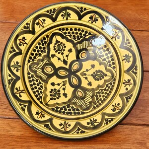 May include: A round, decorative ceramic plate with a yellow background and black floral patterns. The plate features a scalloped edge and a central floral design, with accents of green and orange. The plate is approximately 25 cm in diameter.