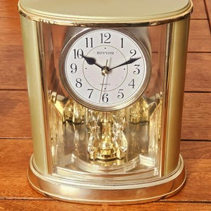 May include: A gold-toned oval mantel clock with a clear glass casing. The white clock face features black Arabic numerals, black hands, and the text RHYTHM QUARTZ. A gold-colored pendulum with clear crystal-like accents hangs below the clock face.