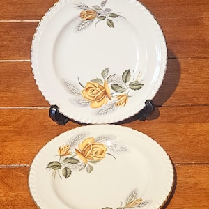 May include: Two white ceramic plates with a gold rim and a raised edge. Each plate features a yellow rose and green leaf design. The plates are displayed on a wooden surface.