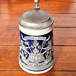 以下が含まれることがあります： 金属製の蓋と球形のつまみが付いた、青と白の陶器製ビアジョッキ。本体にはライオン、小麦、醸造用具の白いレリーフが施されており、ドイツ語で「Hopfen u. Malz」「Gott erhalt's」という文字が刻まれています。