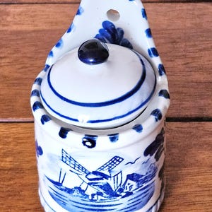 Puede incluir: Un salero de cerámica blanco y azul con tapa y asa. El salero presenta un diseño de molino de viento y detalles azules. La tapa tiene un pomo azul oscuro. El asa tiene un agujero para colgar. El salero mide aproximadamente 10 cm de alto.