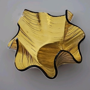 Puede incluir: Un cuenco decorativo de forma libre, de color dorado y ribete negro. El cuenco tiene un diseño ondulado con una superficie texturizada. La forma del cuenco es irregular, con un borde ondulado.