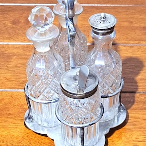 Könnte beinhalten: Ein Vintage-Gewürzset aus Glas mit einem silberfarbenen Metallträger. Das Set enthält vier Flaschen aus diamantgeschliffenem Glas und einen kleinen Behälter mit Klappdeckel. Die Flaschen haben Kristallstopfen und werden von einem dekorativen Metallrahmen gehalten.