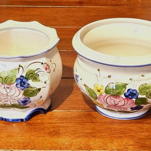 May include: Two off-white ceramic planters featuring hand-painted floral motifs in shades of pink, yellow, blue, and green. The left planter has a scalloped rim and base, while the right planter features a smooth, rounded rim and a circular base.