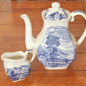 Puede incluir: Un juego de tetera y crema de cerámica blanca con ilustraciones de paisajes azules. La tetera tiene un pico curvo, un asa y una tapa con un diseño floral. La crema tiene un asa pequeña y un patrón de paisaje similar.