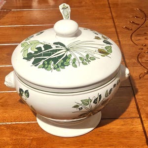 Może przedstawiać: Biała ceramiczna waza do zupy z pokrywką, zdobiona zielonymi i brązowymi wzorami botanicznymi. Waza ma okrągłą podstawę, boczne uchwyty i dekoracyjny uchwyt na pokrywce. Klasyczny element do serwowania zup i gulaszu.