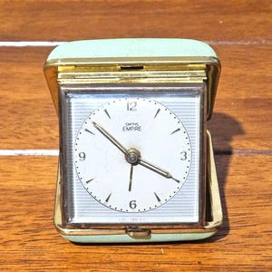 May include: A vintage Smiths Empire travel alarm clock. The clock has a square face with a white dial, black numerals, and gold hands. The clock is housed in a gold-toned metal case with a light green leatherette cover.