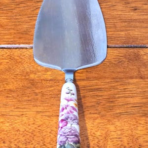 Könnte beinhalten: Ein Kuchenheber aus Edelstahl mit gezahntem Rand und floralem Griff. Der Griff hat eine weiße Basis mit lila und rosa Blumen und grünen Blättern. Der Heber befindet sich vor einem Holzgrund.
