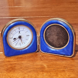 Peut inclure: Un ensemble articulé, bleu et doré, combinant une horloge et un cadre photo. Le cadran de l'horloge est blanc avec des chiffres romains et des aiguilles noirs. Le cadre a un intérieur marron foncé. L'horloge indique "elite".
