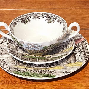 Puede incluir: Un juego de taza y platillo vintage con un diseño de escena pastoral. La taza blanca tiene dos asas y se coloca sobre un platillo a juego y un plato más grande. El juego presenta un paisaje detallado con una paleta de colores marrón, verde y azul.