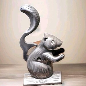May include: A silver-colored squirrel-shaped nutcracker. The nutcracker has a curved tail and is holding a nut. The base is rectangular and has a wood-like texture. The nutcracker is made of metal.