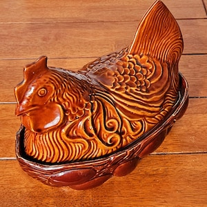May include: A glossy brown ceramic hen-shaped dish with a lid featuring detailed molded feathers and a comb. The base is designed to resemble a woven nest, providing a decorative container for storage or display with a smooth, reflective finish.