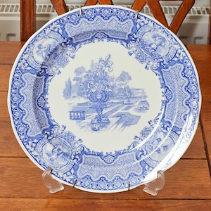 May include: A white and blue decorative plate with a detailed landscape scene in the centre. The plate features a blue floral border and cherub accents. The plate is displayed on a clear stand, showcasing its intricate design.