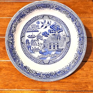 May include: A round, white and blue ceramic plate with a classic Willow pattern design. The plate features a detailed landscape scene with pagodas, trees, and birds. The rim has a decorative border. The plate is set on a wooden surface.