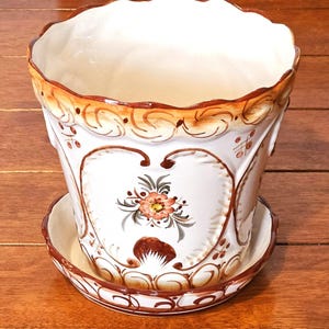 May include: A white ceramic flower pot with a matching saucer. The pot features a scalloped rim and is decorated with brown and orange floral designs. The pot has a heart-shaped design with a flower in the center.