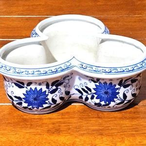 May include: A white ceramic three-section serving dish featuring a blue floral pattern on the exterior. The bowl has a clover-like shape with three rounded compartments and a blue decorative border along the top rim and base of the vessel.