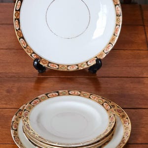 Op de afbeelding: Een set witte porseleinen borden met een decoratieve rand met een herhalend patroon van oranje, zwarte en gouden geometrische vormen. Eén bord staat op een zwarte standaard, andere zijn opgestapeld.