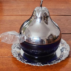 May include: A pear-shaped serving dish with a shiny silver lid and a deep blue base. The dish sits on a decorative silver plate. A small spoon is visible in a plastic wrapper.