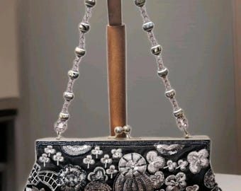 Può includere: Borsa da sera nera con ricami floreali argentati e manico di perline. La borsa ha una forma rettangolare ed è decorata con vari disegni floreali. La borsa è esposta su un supporto di legno.