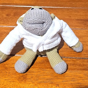 May include: A crocheted stuffed animal featuring a gray head and limbs, olive green torso, and a white terry cloth bathrobe. The toy has a rounded head, long arms, and short legs, with a textured knit surface and a soft fabric robe tied at the waist.