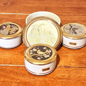 Könnte beinhalten: Eine Sammlung dekorativer Keramikbehälter mit goldenen Rändern und Deckeln. Die Deckel zeigen Vogel- und Blumendesigns in Gold und Schwarz. Die Behälter sind weiß mit einem goldenen Band und dem Text "24KT Gold".