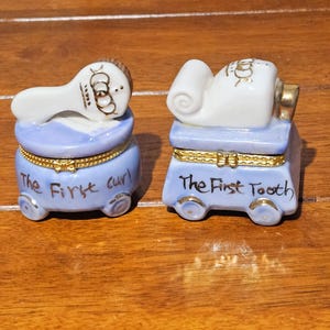 May include: Two porcelain trinket boxes, one shaped like a hairbrush and the other like a tube of toothpaste. Both are light blue with gold accents and the text "The First Curl" and "The First Tooth".