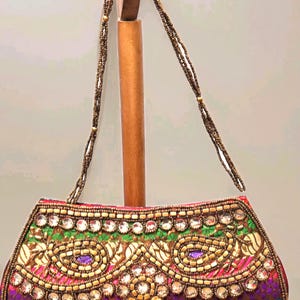 May include: An ornate, rectangular clutch with a beaded handle. The bag features intricate embroidery in gold, green, pink, and purple, accented with clear gemstones. The handle is made of small, brown beads.