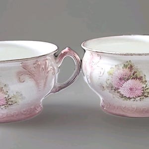 Può includere: Due tazze da tè antiche in porcellana con un motivo floreale. Le tazze sono bianche con dettagli rosa e bordi dorati. Ogni tazza presenta un motivo floreale rosa e un manico curvo.