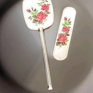 May include: A vintage vanity set with a hand mirror and a comb. Both pieces feature a floral design with pink roses and green leaves on a white background.