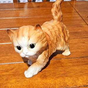 May include: A ceramic figurine of a ginger cat with white markings, black eyes, and a raised tail. The cat is in a walking pose, with its front paw extended. The figurine is on a wooden surface.