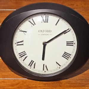 May include: A round wall clock with a black frame and an off-white face. The clock features black Roman numerals and black hands. The text OXFORD STATION is printed in black in the centre of the face. The clock is set to approximately 6:30.