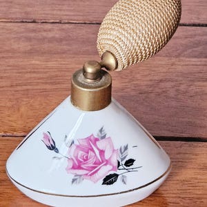 Puede incluir: Botella de perfume blanca vintage con atomizador y detalles dorados. La botella presenta un diseño floral de rosa rosa con hojas negras y grises. El atomizador tiene forma ovalada y texturizada.