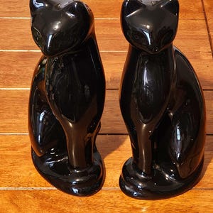 May include: Two glossy black cat figurines. The cats are in a seated position with their heads tilted slightly. The figurines have a smooth, reflective surface and are placed on a wooden surface.