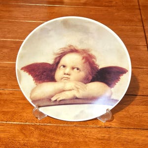 May include: A decorative plate featuring a Renaissance-style cherub with red hair and burgundy wings. The cherub rests its arms on a surface, with a slightly annoyed expression. The plate is white with a light beige background.