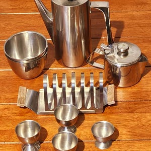 Op de afbeelding: Een roestvrijstalen koffieservies bestaande uit een hoge koffiepot met een ronde dekselknop, een kleinere theepot, een toastrek met houten handvatten, twee in elkaar passende kommen en vier eierdopjes op een voetstuk.