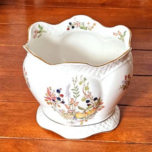 Pode incluir: Uma taça de cerâmica branca com rebordo recortado e acabamento dourado, apresentando uma ilustração botânica colorida de amoras, flores e frutos. A superfície exterior tem um padrão de diamante subtilmente em relevo perto da base.