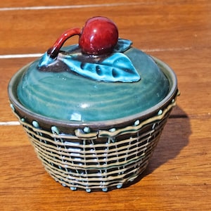 Può includere: Un barattolo in ceramica con coperchio decorato da una ciliegia rosso scuro e una foglia azzurra. La base del barattolo presenta un motivo a cestino intrecciato in tonalità marrone e beige, con accenti azzurri che mettono in risalto il design.