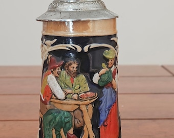 May include: A ceramic beer stein with a detailed relief scene of people at a table, topped with a silver metal lid. The stein has a dark blue background with tan and brown accents, and a decorative base.